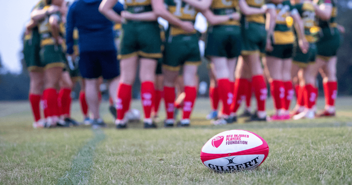 Images shows a group of female rugby players in a huddle, wearing their green and yellow kit with red RFU injured players foundation (IPF) red socks. There's also an RFU IPF ball in the image.